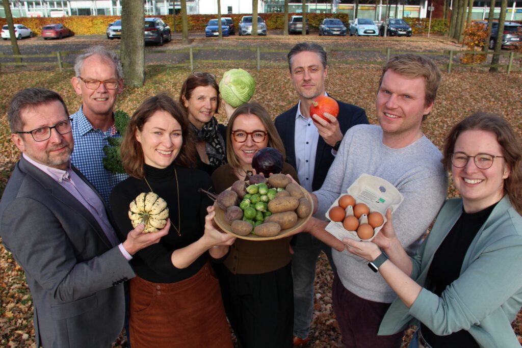 Gruppe von Menschen, die verschiedene Gemüse und Eier in den Händen halten, umgeben von herbstlichem Laub.