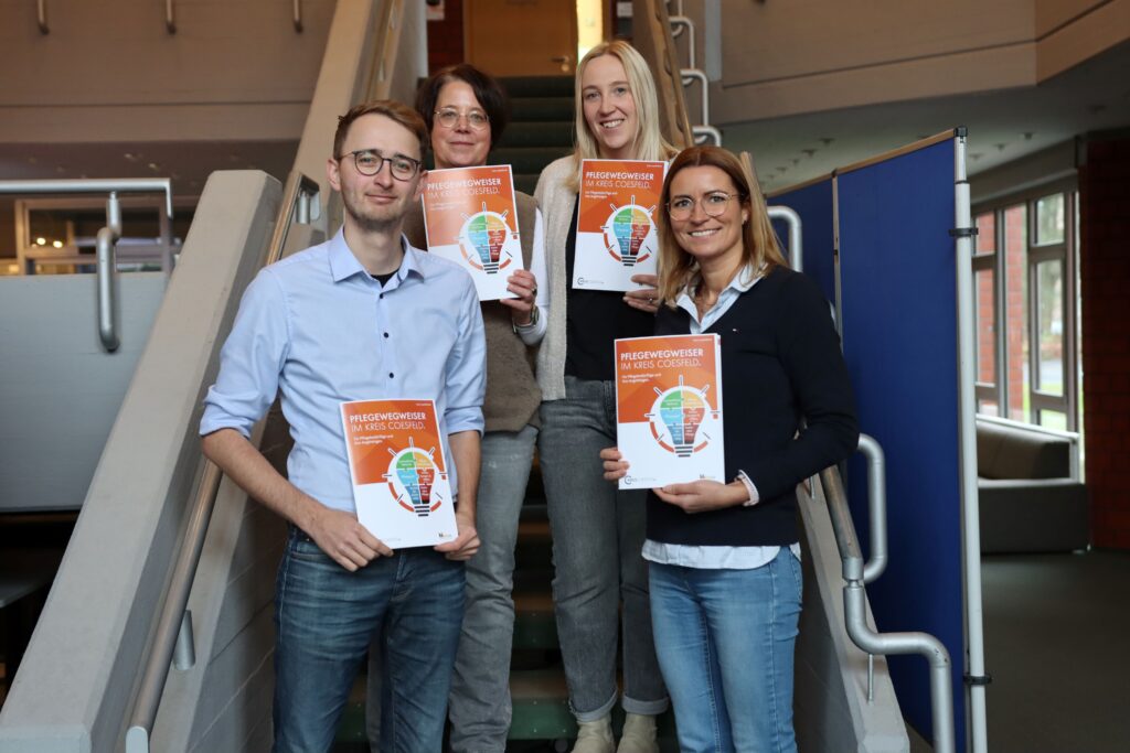 Vier Personen halten Broschüren mit dem Titel 'Pflegewegweiser' in einer Innenumgebung.