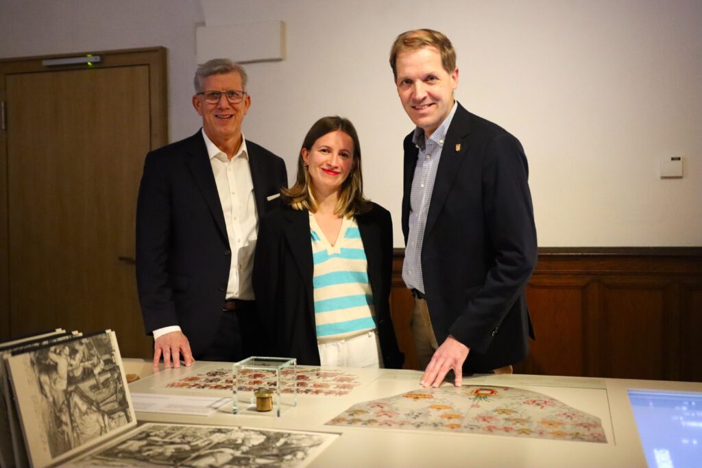 Drei Personen stehen an einem Tisch mit Dokumenten und Zeichnungen in einem Innenraum.