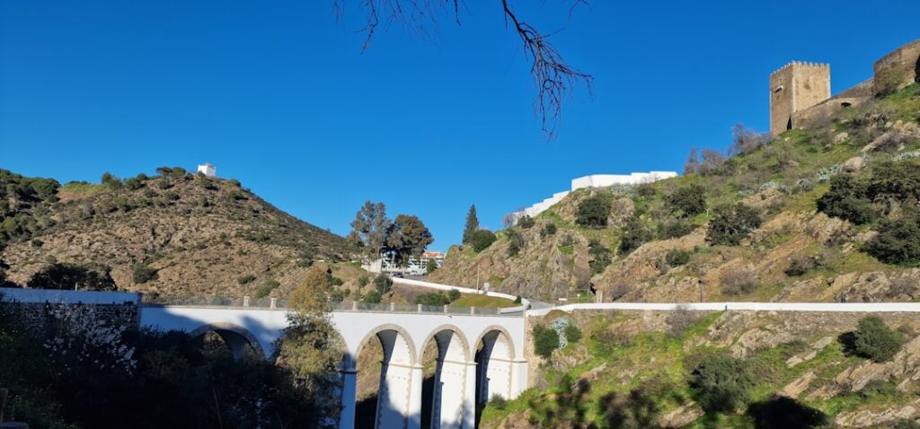 Historische Brücke mit Bögen, umgeben von Hügeln und einer Burg im Hintergrund.