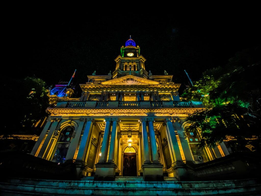 Historisches Gebäude mit beleuchteter Fassade und Uhrturm bei Nacht, umgeben von Bäumen.