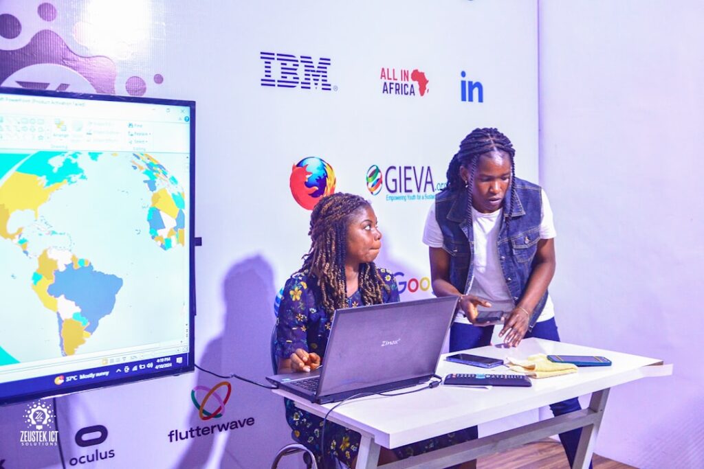 Zwei Personen arbeiten an einem Laptop während einer Präsentation mit Logos von IBM und anderen Unternehmen im Hintergrund.