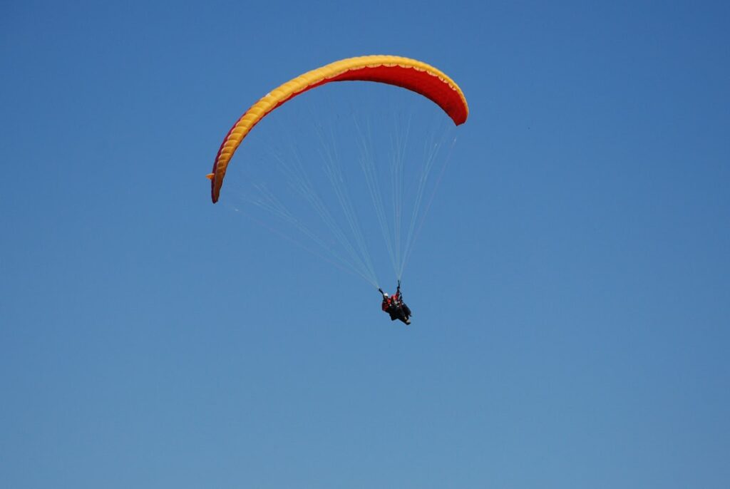 Ein Paraglider mit orangefarbenem Schirm schwebt hoch am blauen Himmel.
