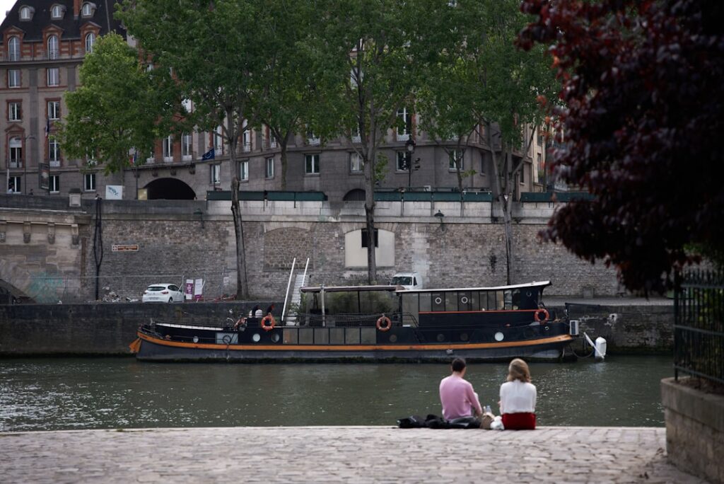 Zwei Personen sitzen am Ufer eines Flusses in einer Stadt, während ein Boot im Wasser liegt.