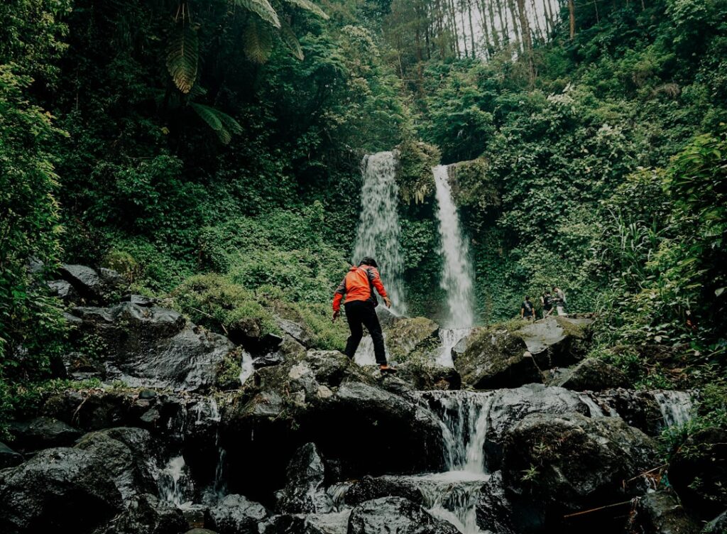 Wanderer in roter Jacke, der über Felsen in einem Waldgebiet mit Wasserfällen klettert.