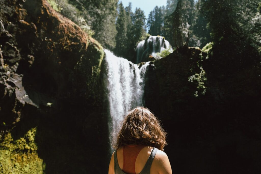 Frau mit lockigem Haar steht vor einem Wasserfall in einer bewaldeten Umgebung.