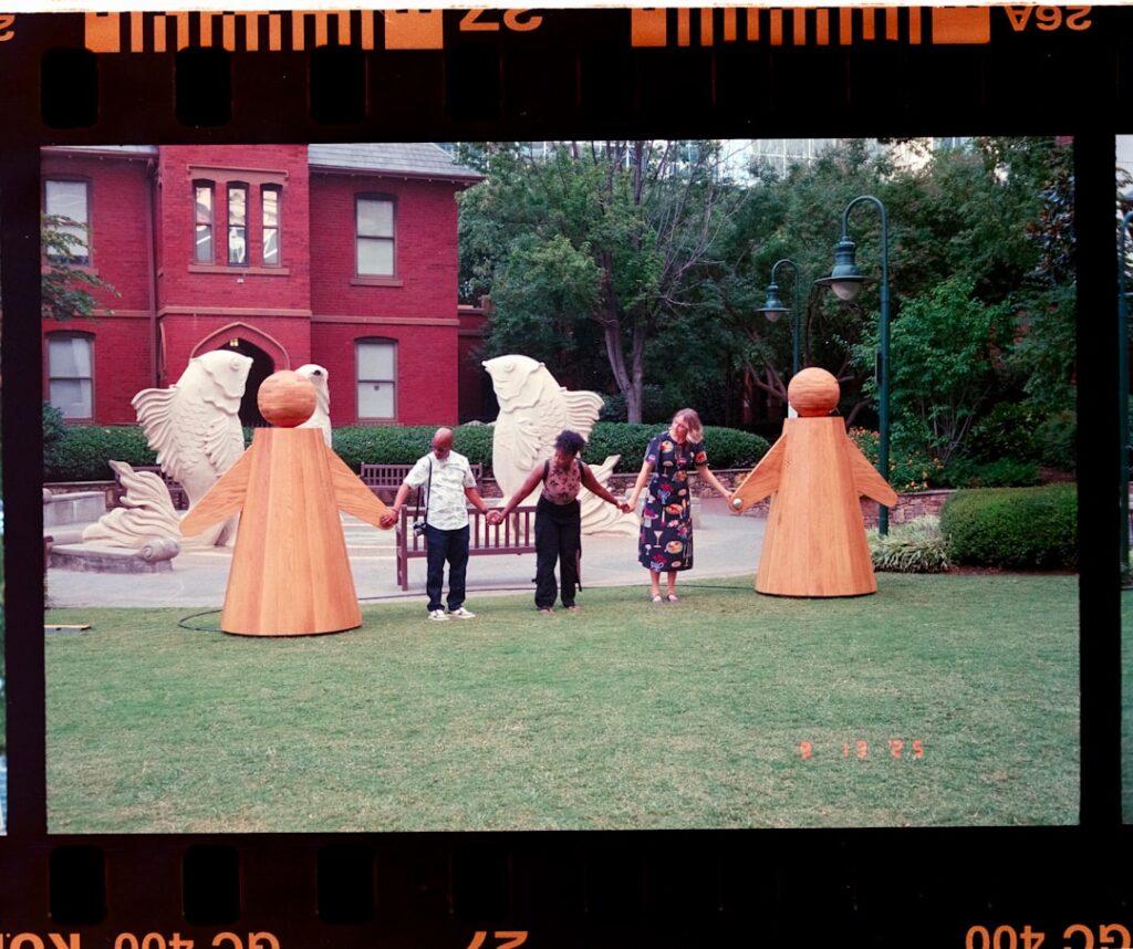 Drei Personen halten sich an den Händen, umgeben von großen Holzskulpturen in Form von Figuren.