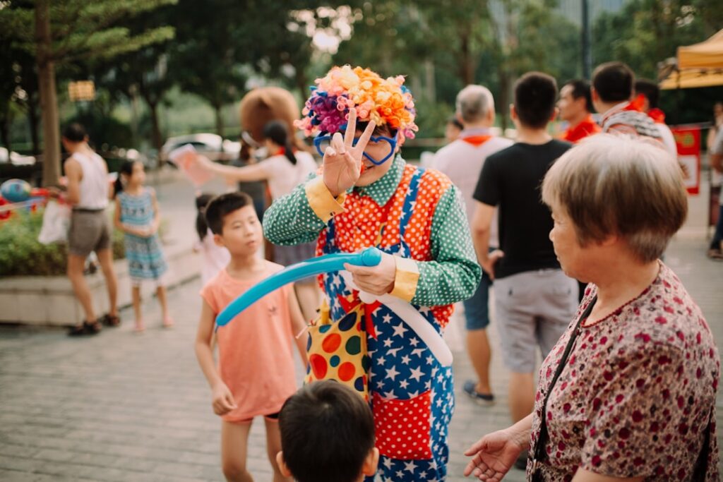 Clown mit buntem Kostüm und Luftballons, der mit Kindern auf einer Veranstaltung interagiert.