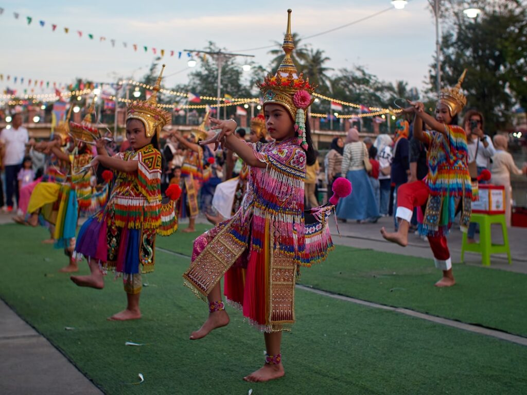 Tanzende Kinder in traditioneller Kleidung bei einer Feier mit bunten Lichtern im Hintergrund.