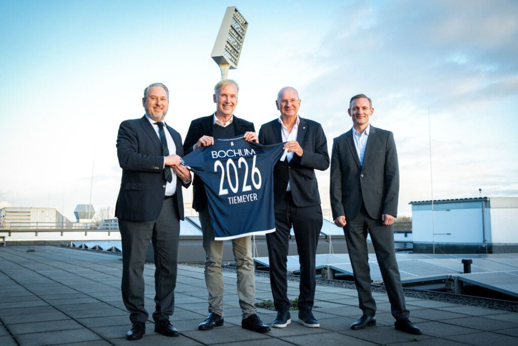 Vier Männer in Anzügen halten ein Trikot mit der Aufschrift 'Bochum 2026' auf einem Dach.