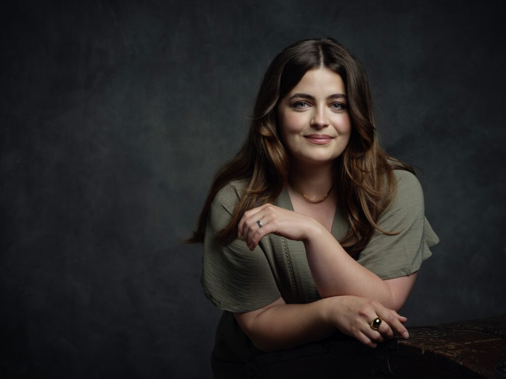 Porträt einer Frau mit langen, braunen Haaren, die in einem grünen Oberteil lächelt und auf einem Tisch lehnt.