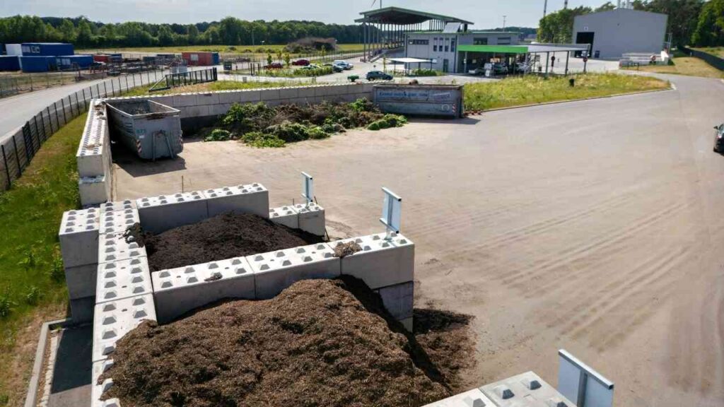 Haufen von Kompostmaterial und Erde auf einem Recyclinggelände mit Containern im Hintergrund.