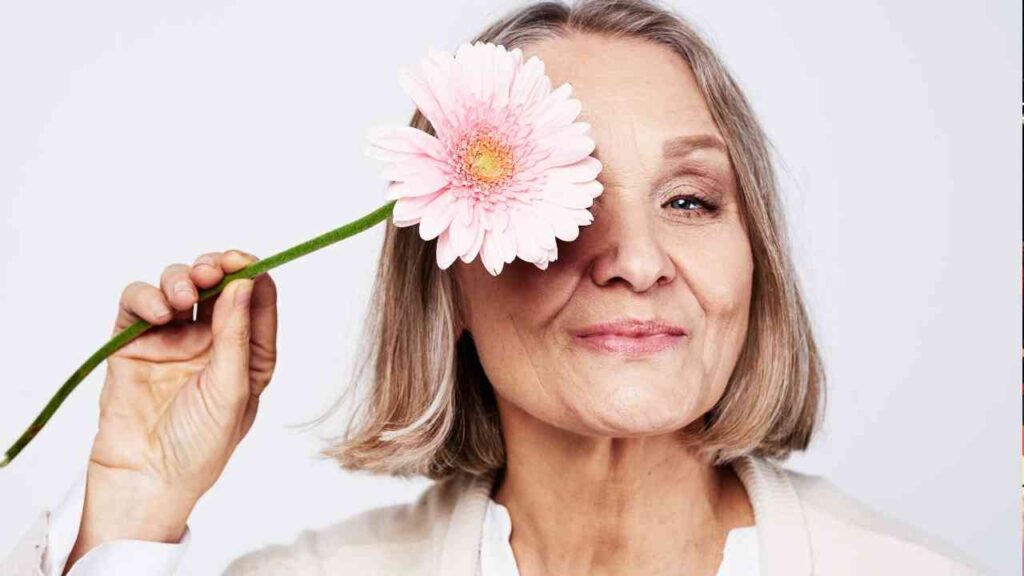 Ältere Frau mit grauen Haaren, die eine pinkfarbene Gerbera-Blume vor ihrem Gesicht hält.