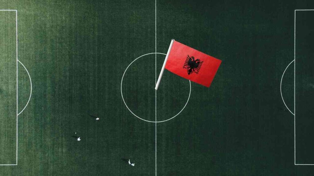 Albanische Flagge auf einem Fußballfeld, mit Spielern im Hintergrund, die sich auf dem Spielfeld bewegen.