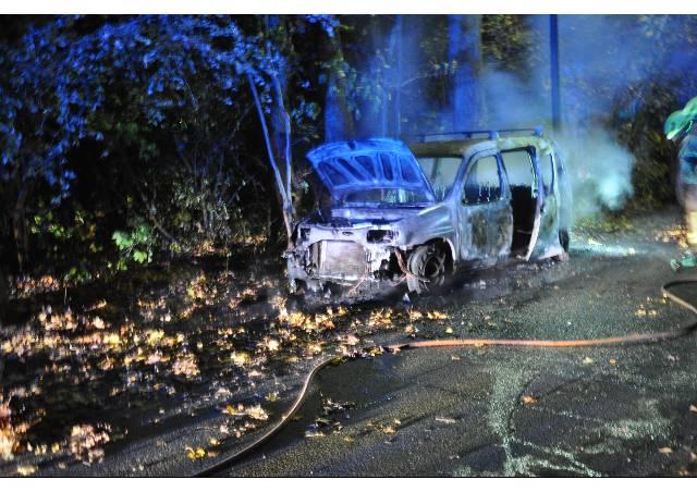 Brennendes Auto mit offenem Motorhaube, umgeben von Rauch und Wasserleitungen in einer nächtlichen Umgebung.