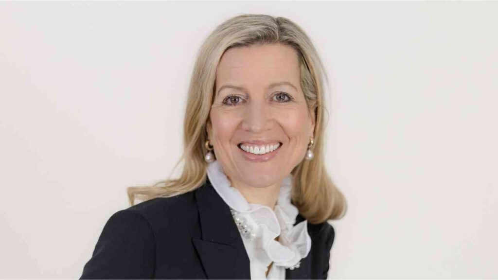 Porträt einer Frau mit blonden Haaren, die in einem schwarzen Blazer und einer weißen Bluse mit Rüschen gekleidet ist.