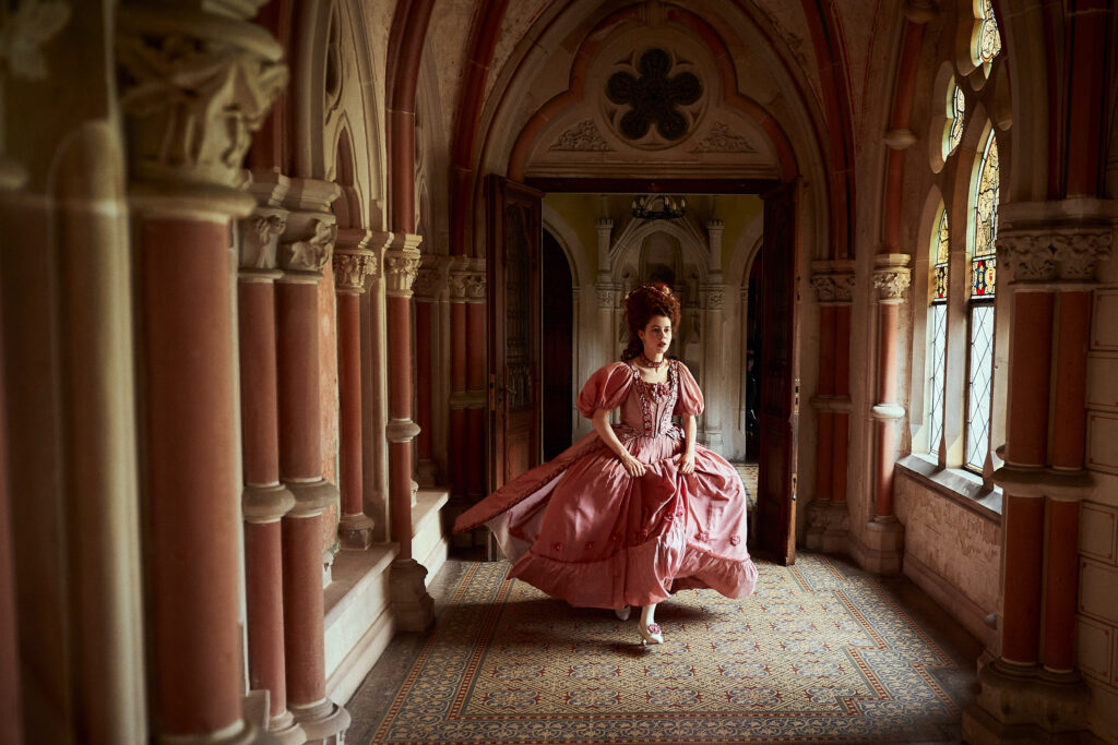 Frau in einem rosa Kleid, die durch einen historisch gestalteten Raum mit Säulen und Fenstern läuft.