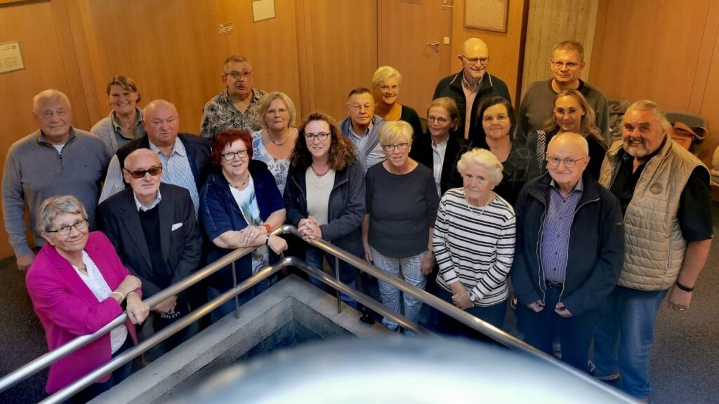 Gruppe von 17 Personen, die in einem Innenraum um ein Geländer versammelt sind, mit verschiedenen Altersgruppen und Geschlechtern.
