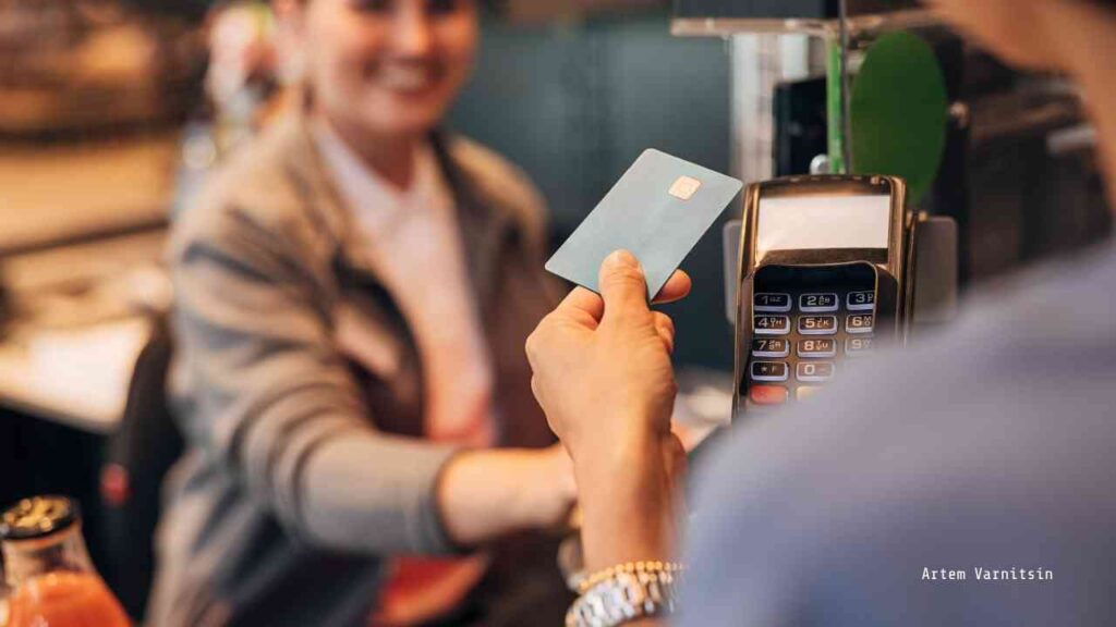Person, die eine blaue Bezahlkarte an ein Zahlungsterminal übergibt, während eine Kassiererin im Hintergrund lächelt.