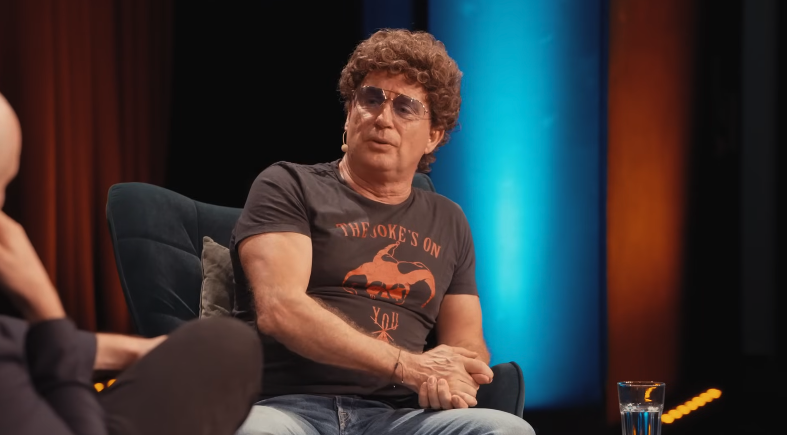 Mann mit lockigen Haaren und T-Shirt sitzt auf einem Sessel während einer Diskussion auf einer Bühne.