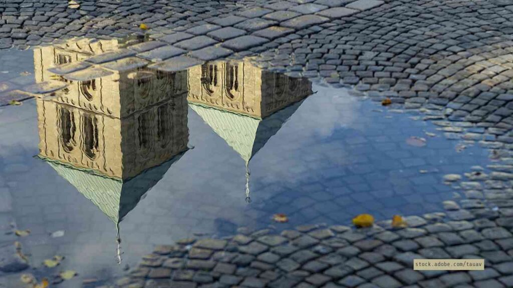 Reflexion der Türme einer historischen Kirche in einer Pfütze auf einem Kopfsteinpflasterweg.