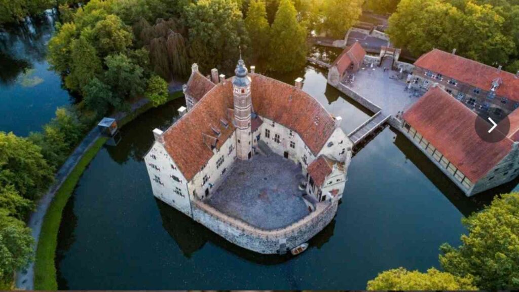 Luftaufnahme von Burg Vischering, umgeben von Wasser und Bäumen, mit roten Dächern und historischer Architektur.