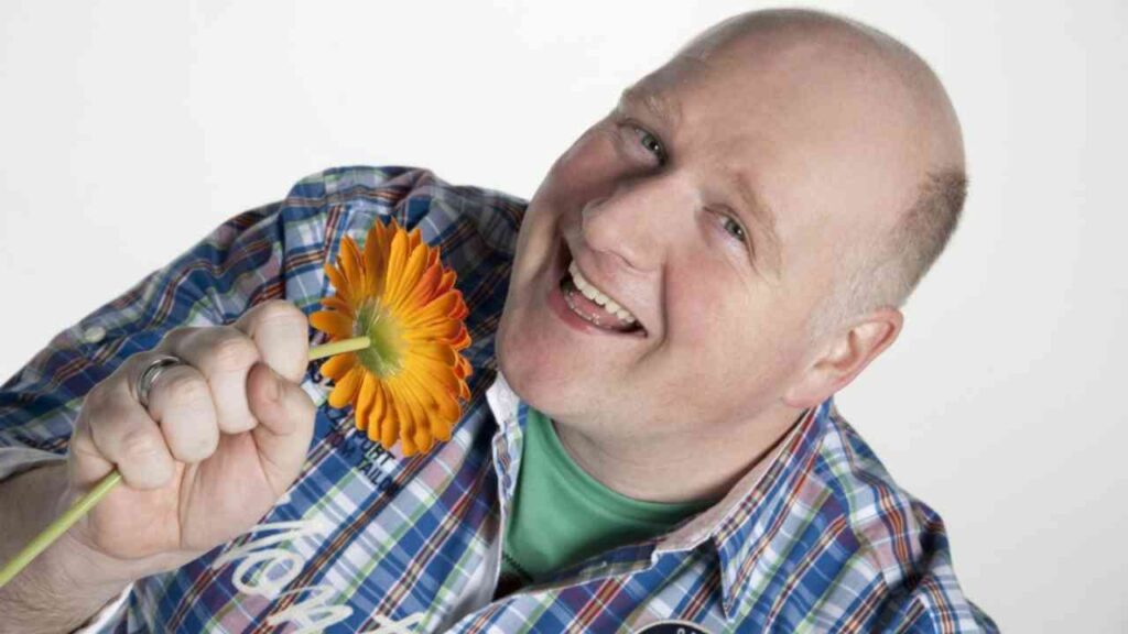 Mann mit einem orangefarbenen Blütenstängel in der Hand, lächelnd und in einem karierten Hemd.