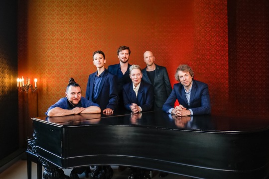 Sechs Musiker in Anzügen posieren um ein schwarzes Klavier in einem stilvollen Raum.