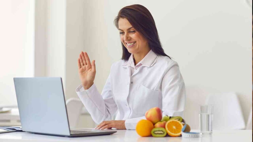 Frau in einem weißen Kittel, die vor einem Laptop sitzt und mit einer Hand winkt, während Obst auf dem Tisch liegt.