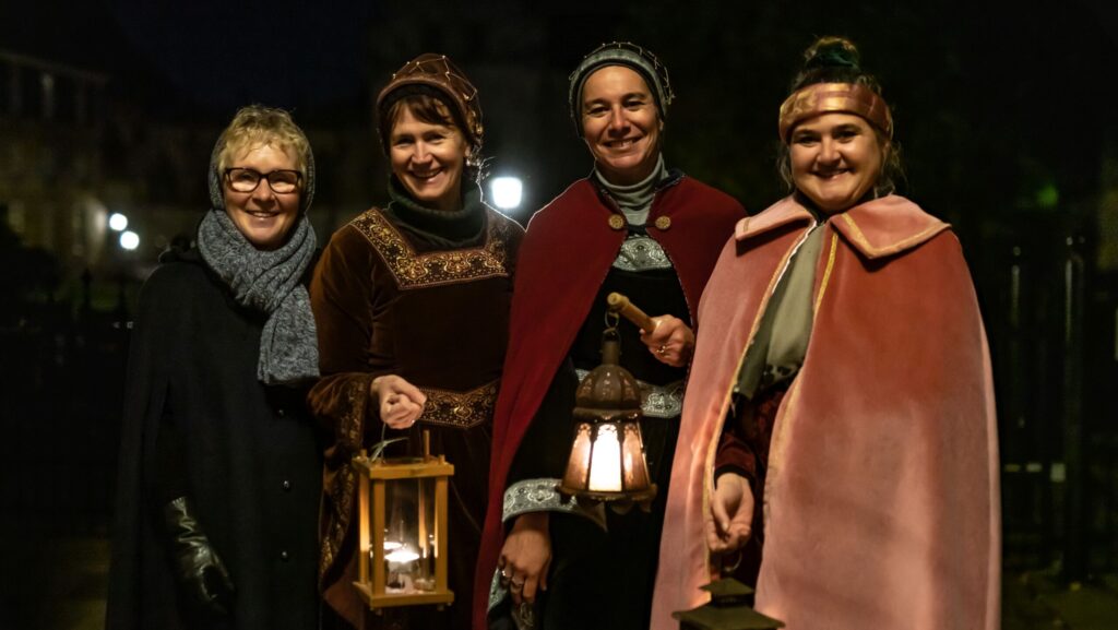 Vier Frauen in historischen Kostümen halten Laternen und posieren für ein Foto bei Nacht.