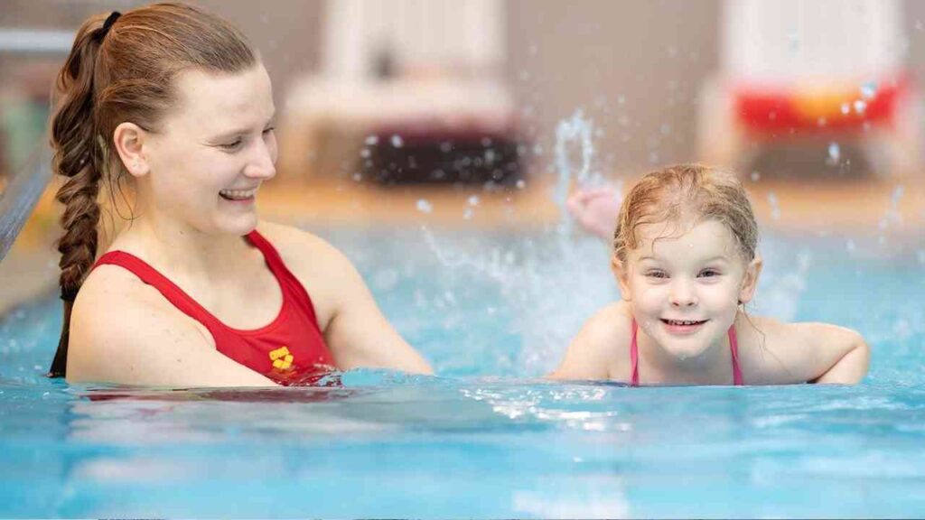 Eine Schwimmtrainerin mit Zopf, die mit einem lächelnden Mädchen im Wasser spielt.