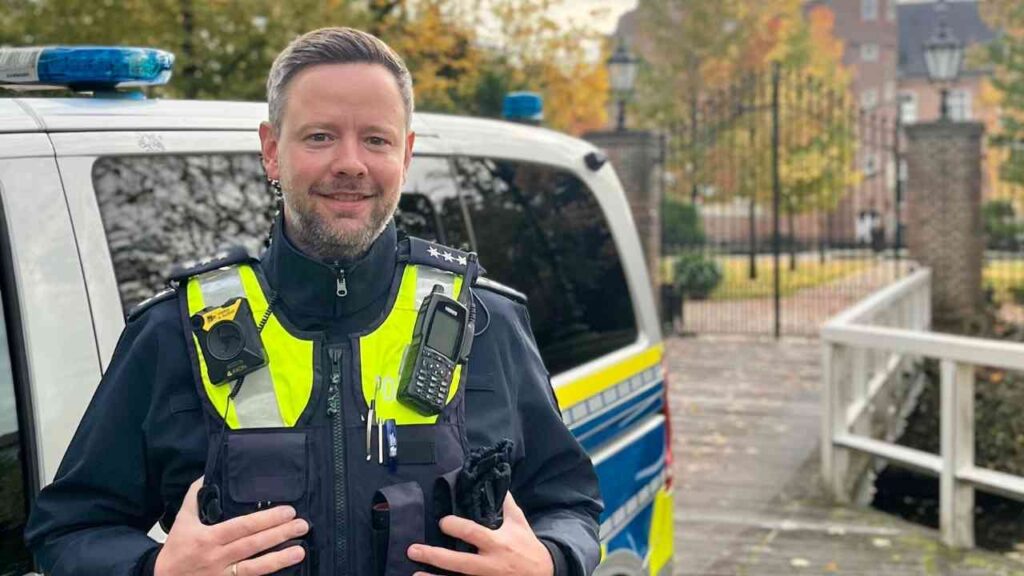 Polizist in Uniform steht vor einem Polizeifahrzeug mit reflektierenden Elementen und einem Lächeln.