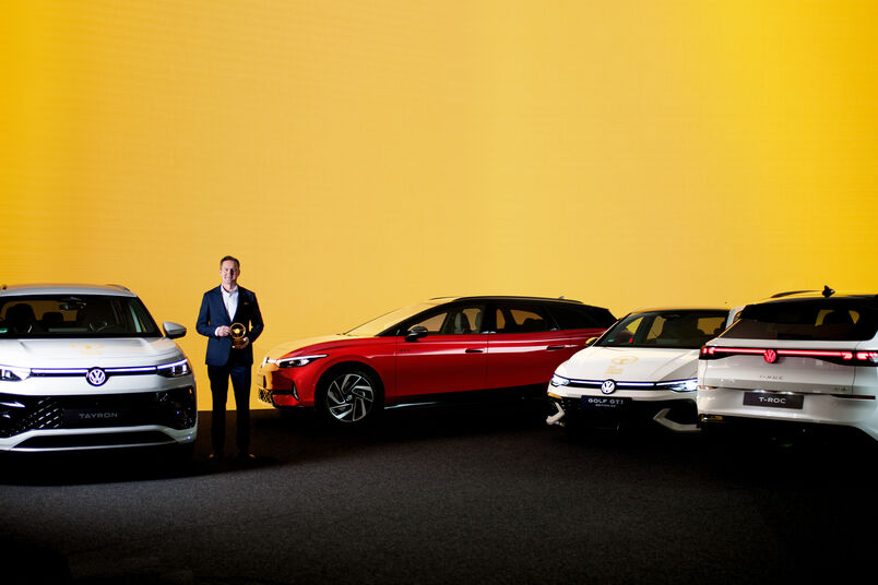 Mann mit Auszeichnung steht zwischen mehreren Volkswagen Fahrzeugen, darunter ID.4, ID. Buzz, T-Roc und Tayron vor gelbem Hintergrund.