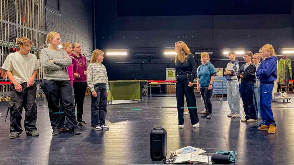 Gruppe von Jugendlichen und einer Lehrerin während einer Theaterprobe in einem dunklen Raum mit Bühnenbeleuchtung.