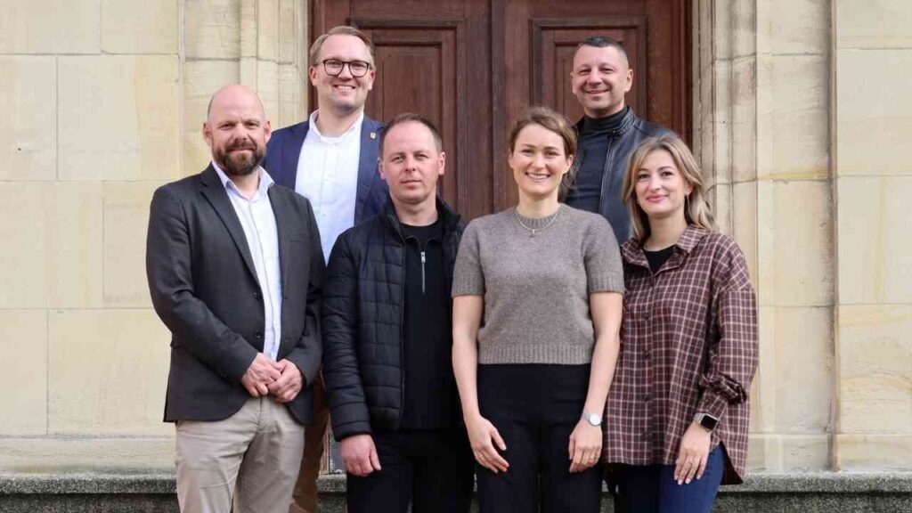 Sechs Personen posieren vor einer Holztür, darunter Männer in Anzügen und Frauen in lässiger Kleidung.