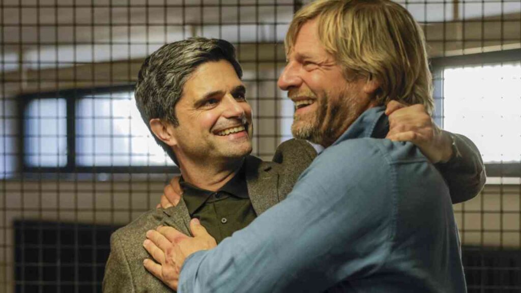 Zwei Männer umarmen sich lächelnd in einem Innenraum mit Gitterfenstern.