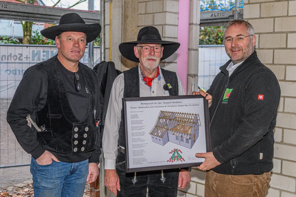 Drei Männer in traditioneller Kleidung präsentieren eine Bauzeichnung eines Holzhauses in einem Innenraum.