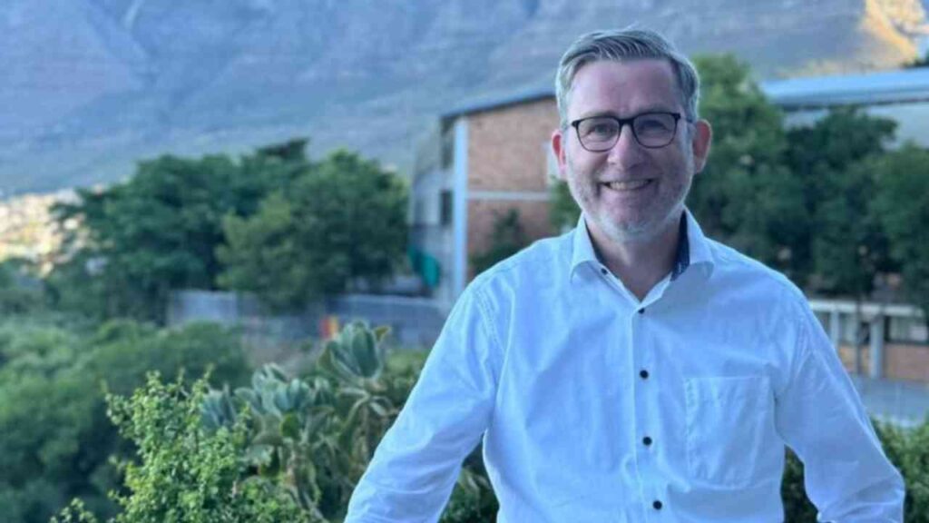 Mann mit Brille und weißem Hemd steht im Freien vor einer grünen Landschaft und einem Gebäude.