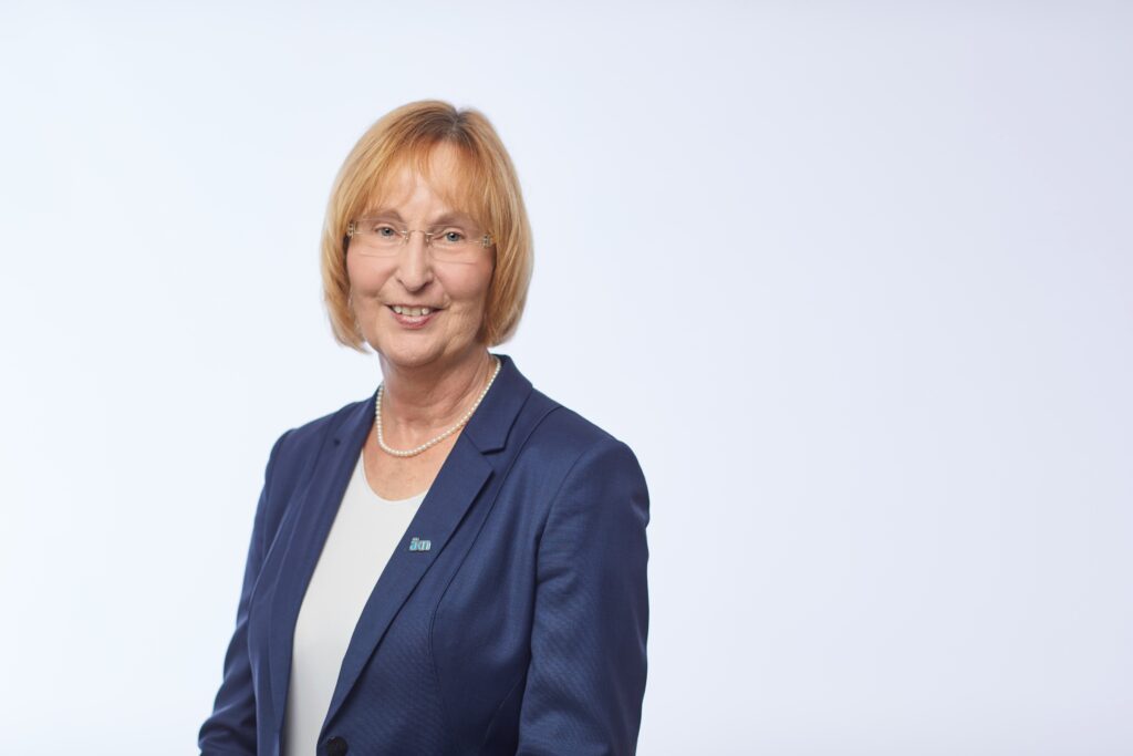 Frau mit blonden Haaren in einem blauen Blazer, lächelnd vor einem hellen Hintergrund.