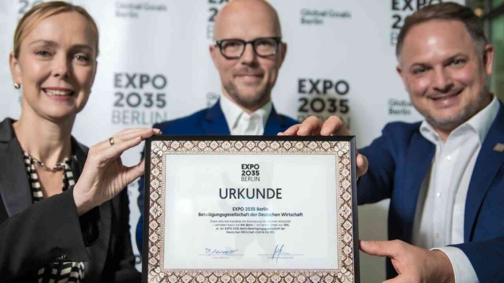 Drei Personen halten eine Urkunde für die EXPO 2035 in Berlin, mit dem Titel 'URKUNDE' deutlich sichtbar.