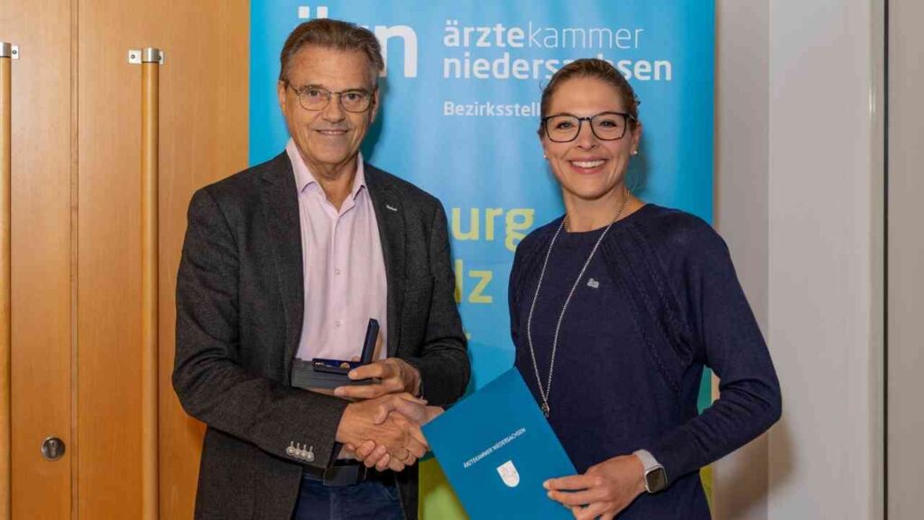 Zwei Personen schütteln sich die Hände, während eine Auszeichnung überreicht wird, im Hintergrund ein Banner der Ärztekammer Niedersachsen.