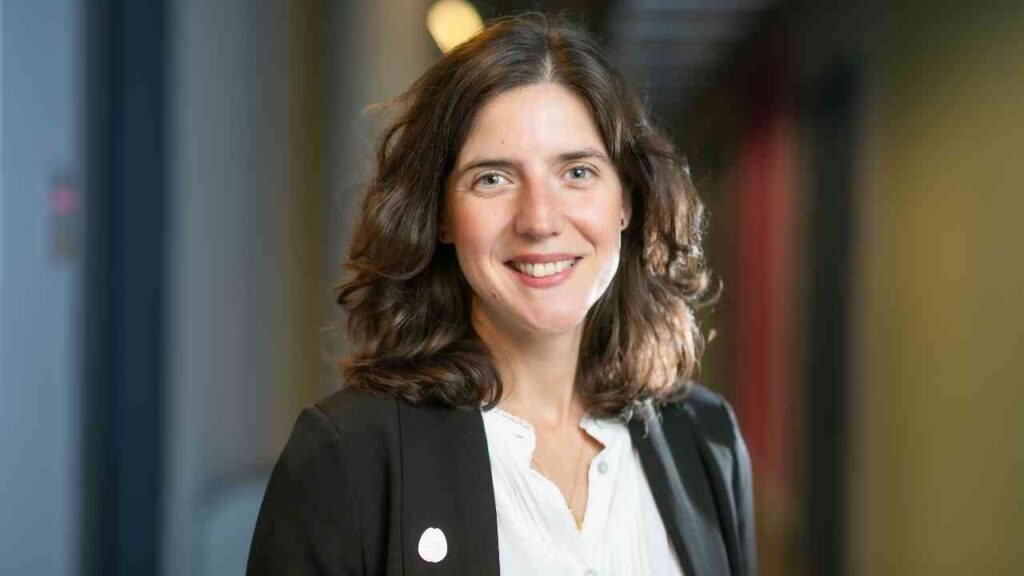 Porträt einer Frau mit lockigem Haar, die in einem Bürogebäude lächelt und einen schwarzen Blazer trägt.