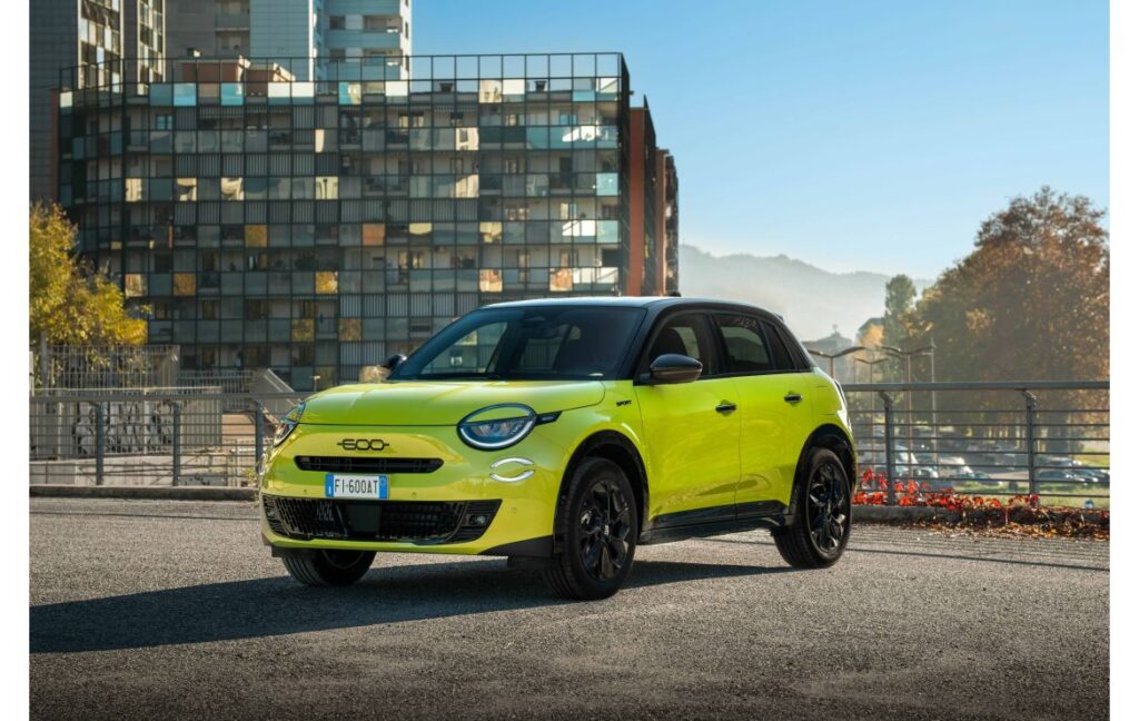 Gelber Fiat 600 in einer urbanen Umgebung mit modernen Gebäuden im Hintergrund.