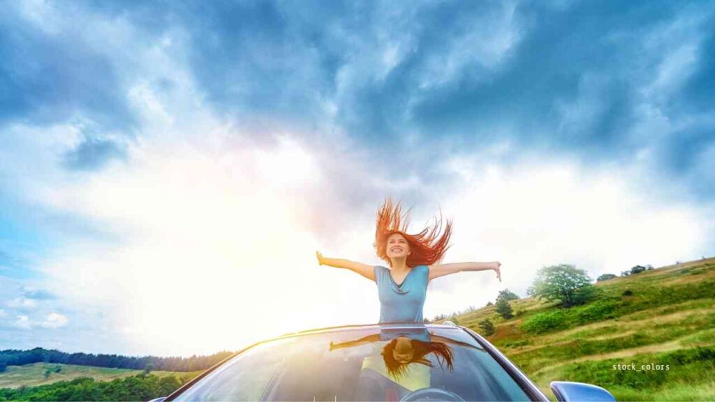 Junge Frau mit langen Haaren, die auf dem Dach eines Autos steht und die Arme ausbreitet, während sie in die Landschaft blickt.