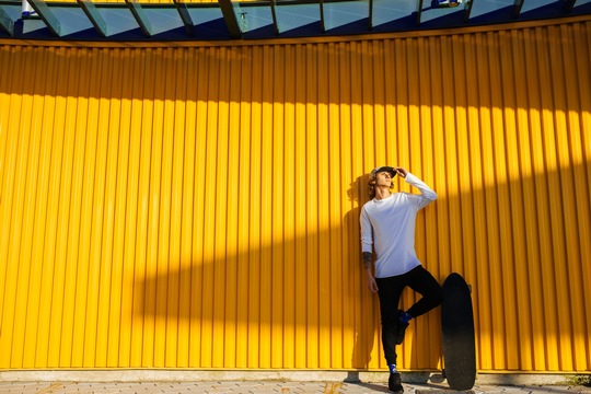 Junger Mann in einem weißen Shirt lehnt an einer gelben Wand und hält ein Skateboard.