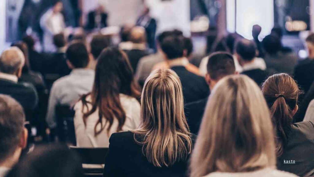Menschen in einem Konferenzraum, die einer Präsentation oder Diskussion auf einer Bühne folgen.