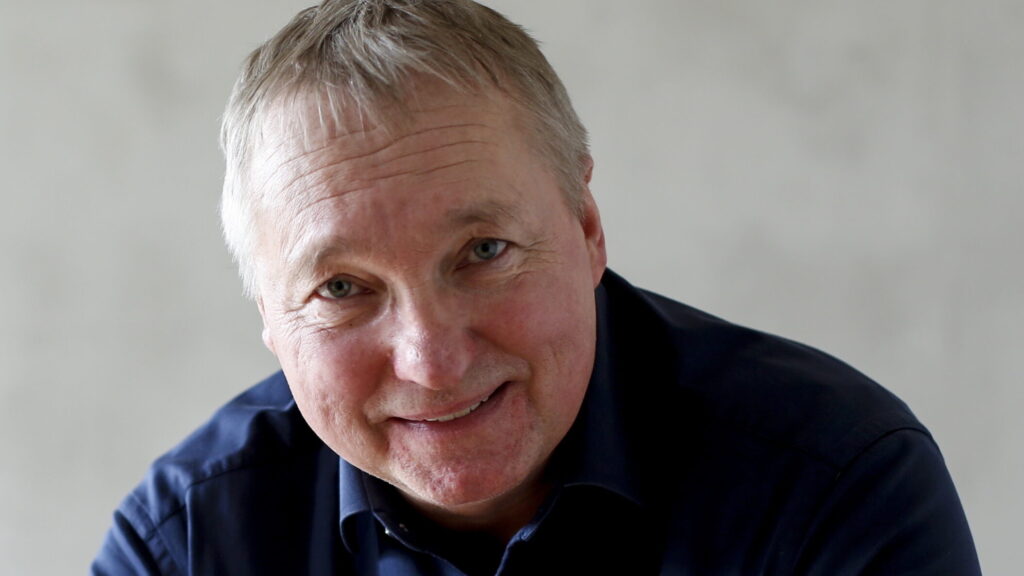 Ein Mann mit kurzen, grauen Haaren und einem freundlichen Lächeln, der ein dunkelblaues Hemd trägt.