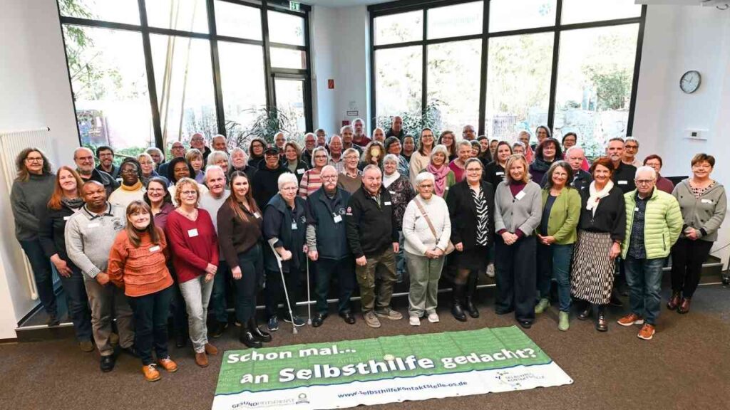 Gruppenfoto von Teilnehmern einer Selbsthilfeveranstaltung in einem Innenraum mit Banner im Vordergrund.