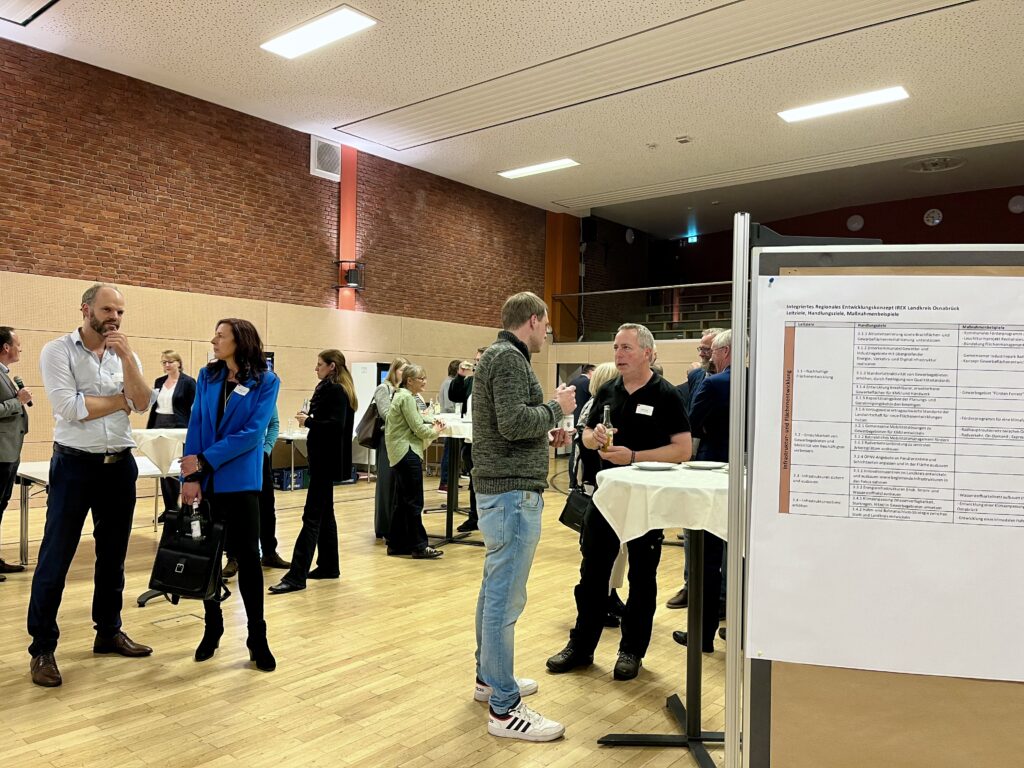 Menschen in einem Raum, die an Stehtischen stehen und miteinander sprechen, während eine Präsentation im Hintergrund sichtbar ist.