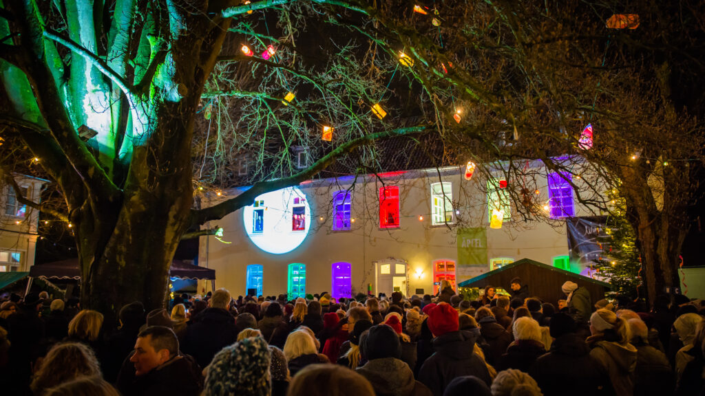 Bunte Fensterbeleuchtung an einem Gebäude mit Menschenmenge bei Nacht, umgeben von einem Baum mit Lichtern.